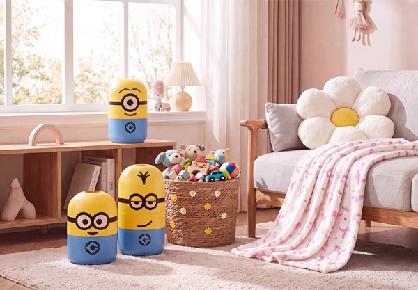 Kinderzimmer mit Minions-Aufbewahrungsboxen Drei gelb-blaue Aufbewahrungsbehälter im Minions-Design stehen in einem hell eingerichteten Kinderzimmer neben einem Korb mit Spielzeug. Auf einem Sessel liegen ein Blumenkissen und eine rosa gemusterte Decke.