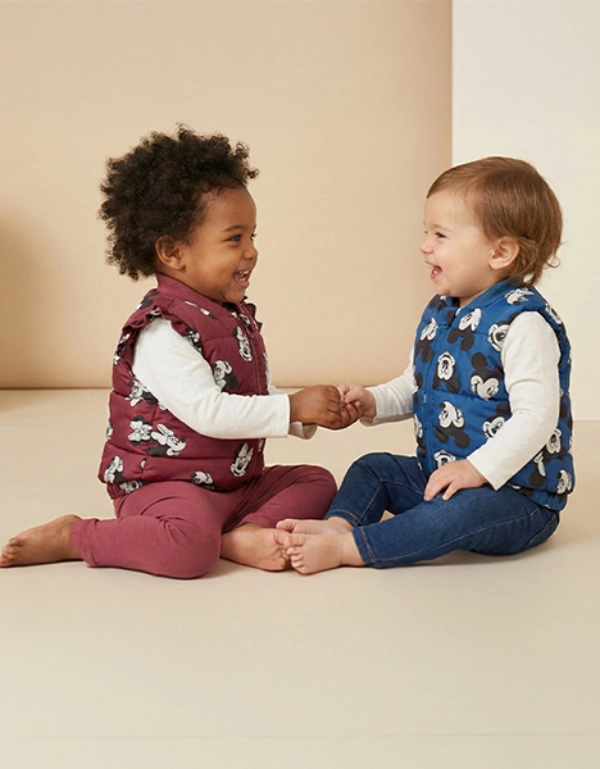 Zwei Kleinkinder sitzen vor einem hellen Hintergrund, tragen bunte Westen, halten sich an den Händen und lächeln sich gegenseitig an.