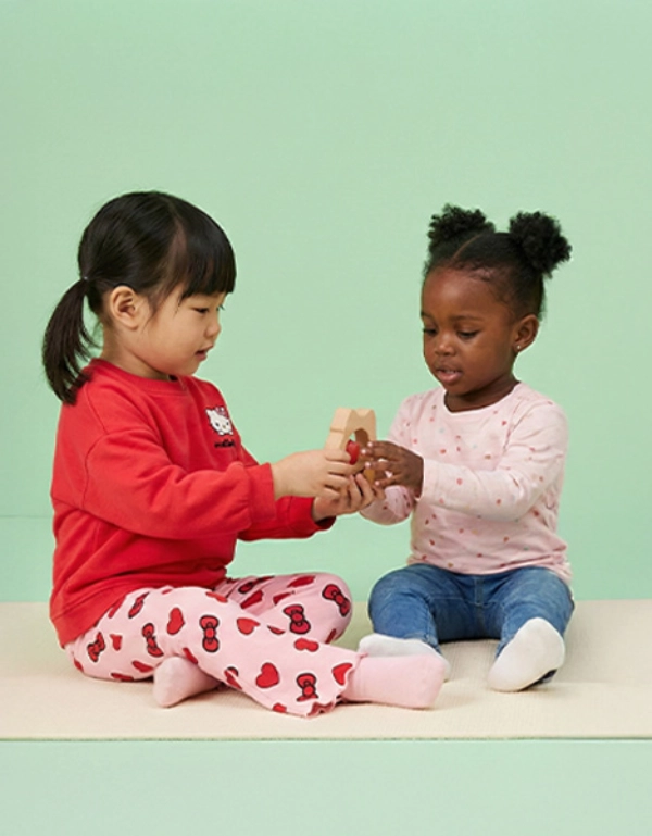 Zwei kleine Kinder sitzen auf einer weichen Unterlage und spielen fröhlich miteinander, während sie ein buntes Spielzeug vor einem pastellgrünen Hintergrund halten.