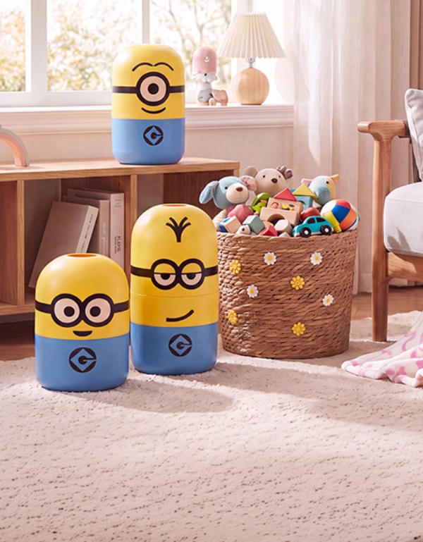 Drei gelb-blaue Aufbewahrungsbehälter im Minions-Design stehen in einem hell eingerichteten Kinderzimmer neben einem Korb mit Spielzeug. Auf einem Sessel liegen ein Blumenkissen und eine rosa gemusterte Decke.