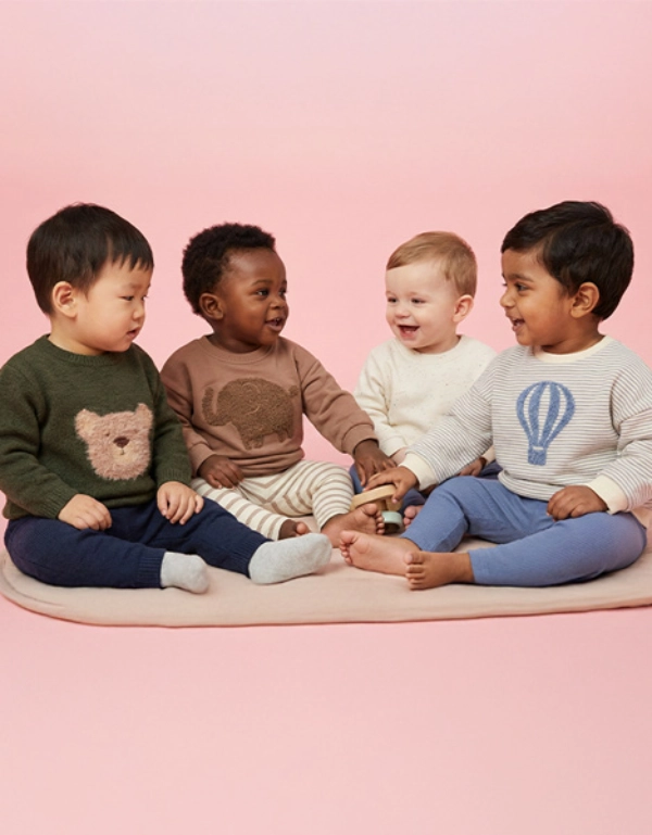Vier Kinder sitzen auf einem Kissen vor einem rosa Hintergrund und tragen bunte Pullover mit verspielten Motiven.