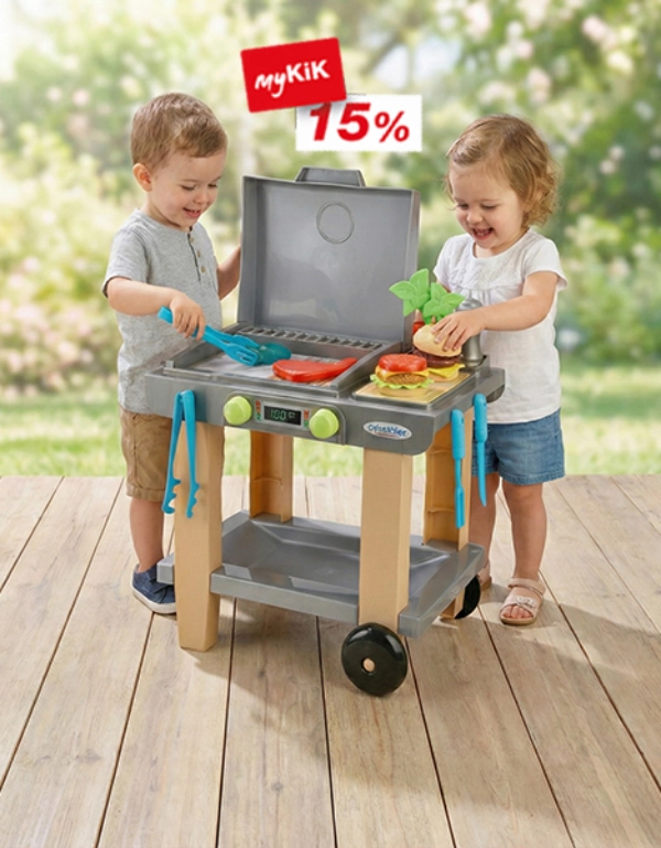 Zwei Kinder spielen mit einem Spielzeuggrill, bunten Utensilien und Spielzeuglebensmitteln auf einer von Grün umgebenen Holzterrasse.