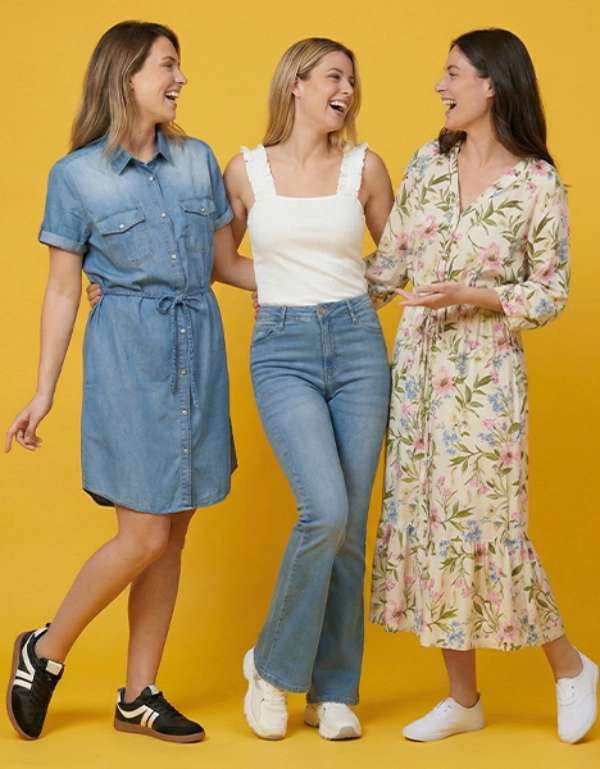 Drei Frauen stehen zusammen vor einem leuchtend gelben Hintergrund und tragen jeweils stilvolle Outfits: ein Jeanskleid, ein lässiges Oberteil mit Jeans und ein geblümtes Maxikleid.