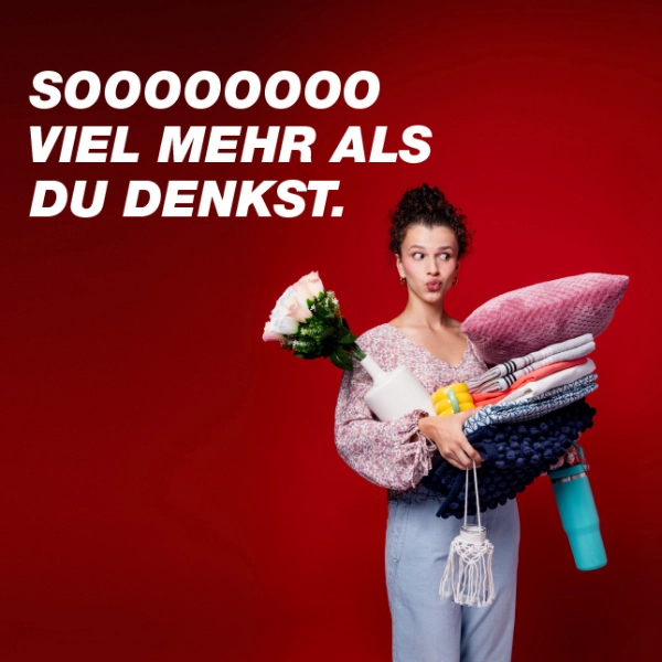 Eine Person hält mehrere bunte Gegenstände, darunter Decken, eine Wasserflasche und Blumen, vor einem leuchtend roten Hintergrund.
