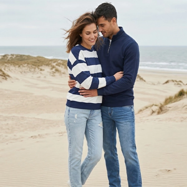 Ein Paar in lässiger Kleidung umarmt sich an einem Sandstrand mit Dünen und Meereswellen unter einem bewölkten Himmel.