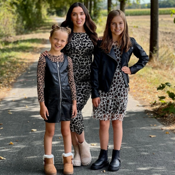 Drei Kinder posieren auf einem mit Blättern übersäten Weg, gekleidet in stilvolle Outfits mit Leopardenmuster, umgeben von Bäumen und einem klaren Himmel über ihnen.