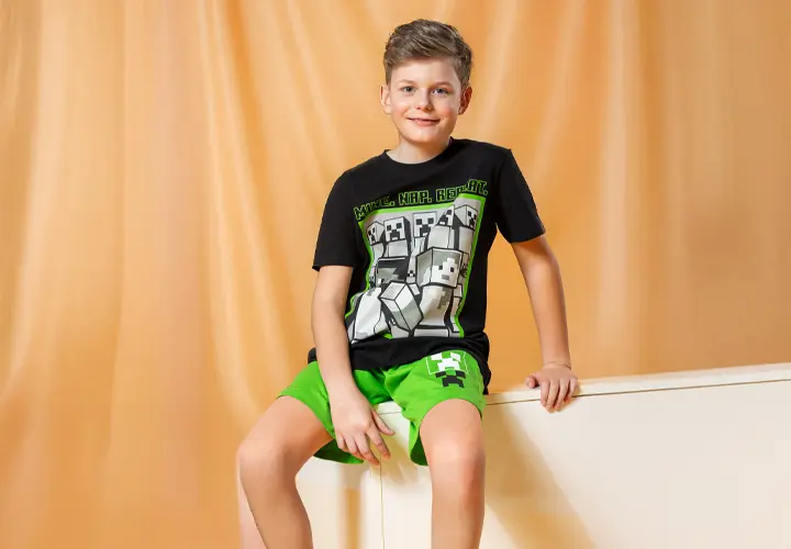 Minecraft Kleidung für Kinder Junge sitzt in schwarzem Minecraft T-Shirt und grüner Minecraft Hose vor gelber Gardine.