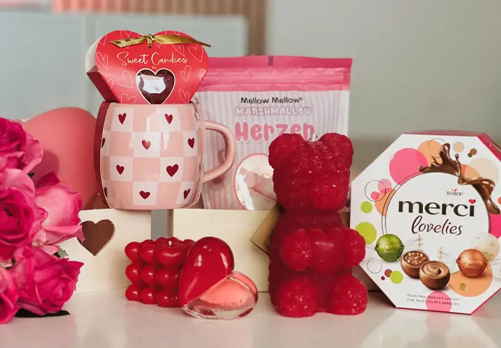 Zeig deine Liebe! Ein festliches Arrangement von Valentinstagsgeschenken: eine Tasse mit Herzmuster, Süßigkeiten, ein roter Gummibär und eine Pralinenschachtel, dekoriert mit Rosen.