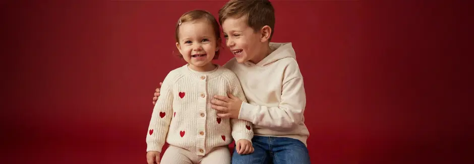 Zwei Kinder sitzen nebeneinander, tragen gemütliche Kleidung und lächeln vor einem satten roten Hintergrund. Das Mädchen trägt eine cremefarbene Strickjacke mit roten Herzen.