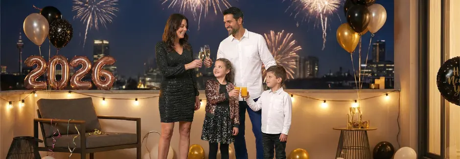 Eine Familie feiert mit Getränken auf einem Balkon, während hinter ihnen ein Feuerwerk den Nachthimmel erhellt.