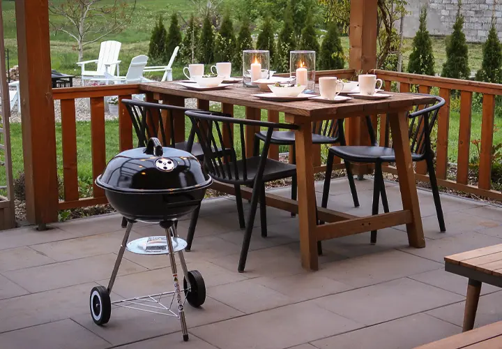 Ein gemütlicher Essbereich im Freien mit einem Holztisch mit Kerzen, Stühlen und einem kleinen schwarzen Grill daneben, inmitten eines grünen Gartens.