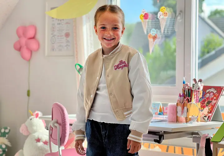 Ein Kind in einer stylischen beige-weißen Collegejacke steht in einem bunten, fröhlichen Raum voller Bastelmaterialien und Dekorationen.