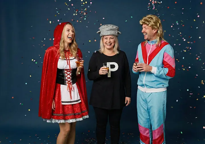 Drei Personen in bunten Kostümen feiern mit Getränken, umgeben von lebendigem Konfetti vor einem blauen Hintergrund.