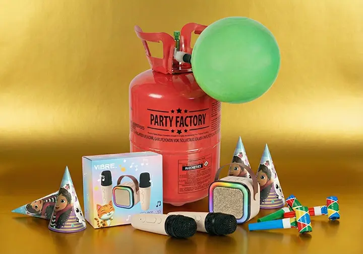 Ein buntes Party-Setup mit einem Heliumtank, Ballons, Partyhüten, Musikmikrofonen und einer lebhaften Geschenkschachtel vor einem goldenen Hintergrund.