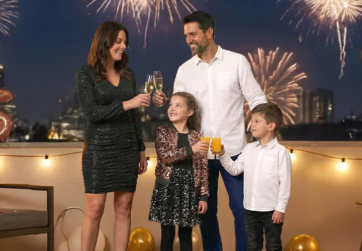 Eine Familie feiert Silvester auf einem Balkon, der mit Luftballons und Lichtern geschmückt ist, während Feuerwerke den Nachthimmel erleuchten.