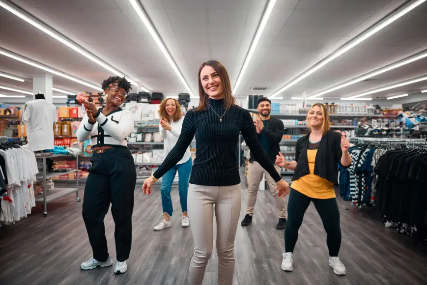 Gruppe von Personen tanzt und klatscht in einem Bekleidungsgeschäft, umgeben von Regalen mit Kleidung.