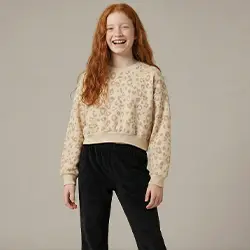 Mädchen trägt einen beigen Strickpullover und schwarze Hose vor hellem Hintergrund.