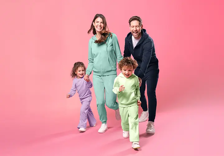 Eine vierköpfige Familie in farbenfrohen, aufeinander abgestimmten Outfits läuft fröhlich Hand in Hand vor einem zartrosa Hintergrund.