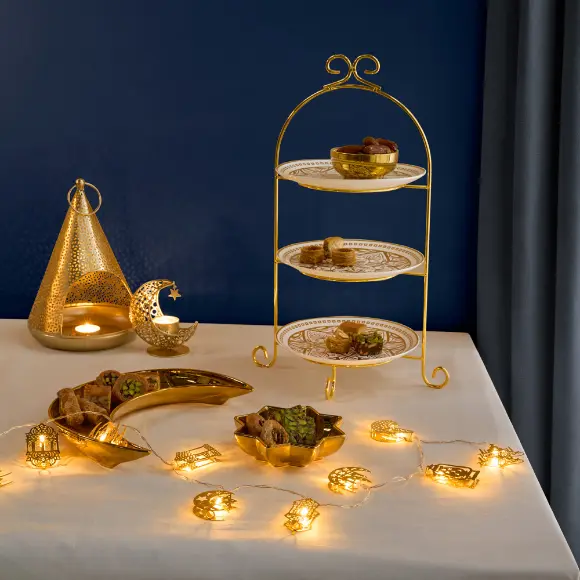 Ein wunderschön arrangierter Desserttisch mit einem goldenen dreistöckigen Ständer mit Gebäck, Laternen und funkelnden Lichterketten vor einem tiefblauen Hintergrund. Ein wunderschön arrangierter Desserttisch mit einem goldenen dreistöckigen Ständer mit Gebäck, Laternen und funkelnden Lichterketten vor einem tiefblauen Hintergrund.