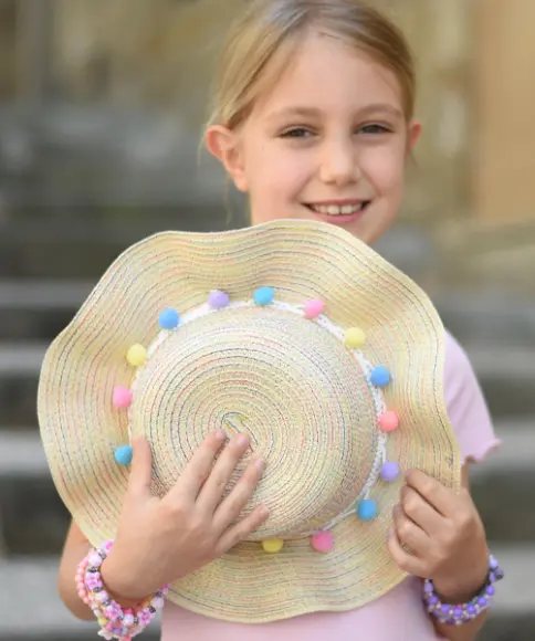 Ein Mädchen hält lächelnd einen cremefarbenen Strohhut mit kleinen bunten Pompons in der Hand