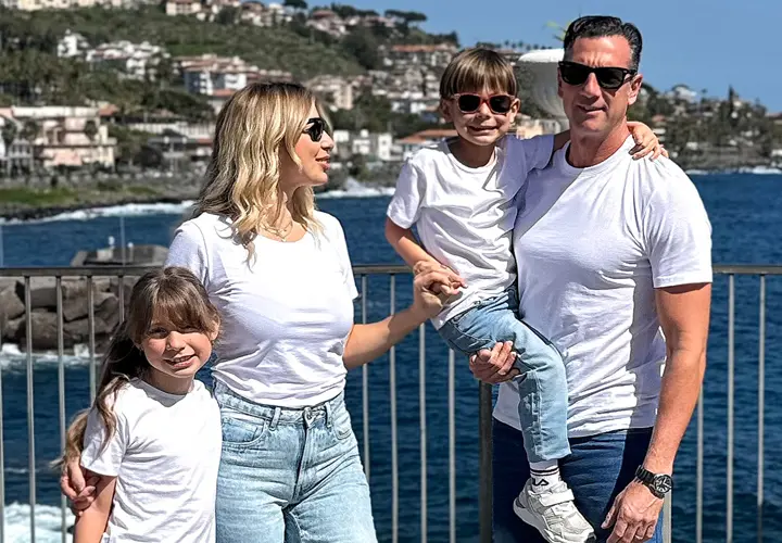 Eine vierköpfige Familie posiert gemeinsam am Meer, trägt passende weiße T-Shirts und Jeans, mit einer malerischen Kulisse aus Hügeln und Wasser im Hintergrund.