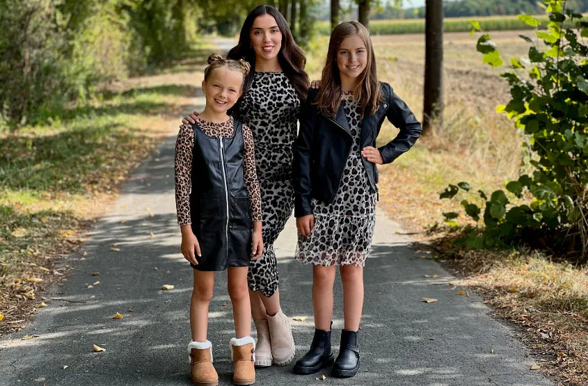 Drei Kinder posieren auf einem mit Blättern übersäten Weg, gekleidet in stilvolle Outfits mit Leopardenmuster, umgeben von Bäumen und einem klaren Himmel über ihnen.