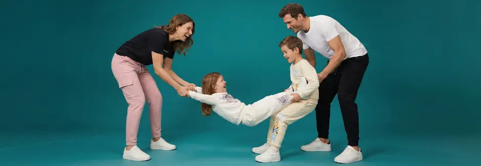 Eine Familie interagiert spielerisch miteinander und hält ein Kind zwischen zwei Erwachsenen; alle sind lässig gekleidet vor einem türkisfarbenen Hintergrund.