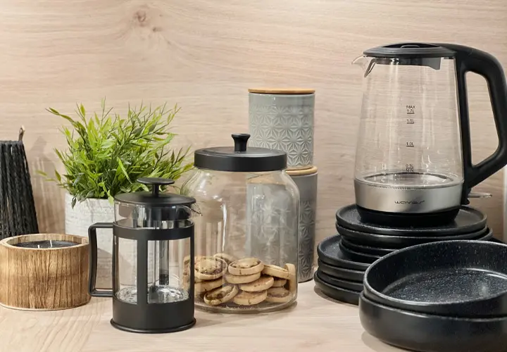 Eine gemütliche Küchenszene mit einem Glaskessel, einer French Press, einer Keksdose, Pflanzendekor und verschiedenem Geschirr vor einem Holzhintergrund