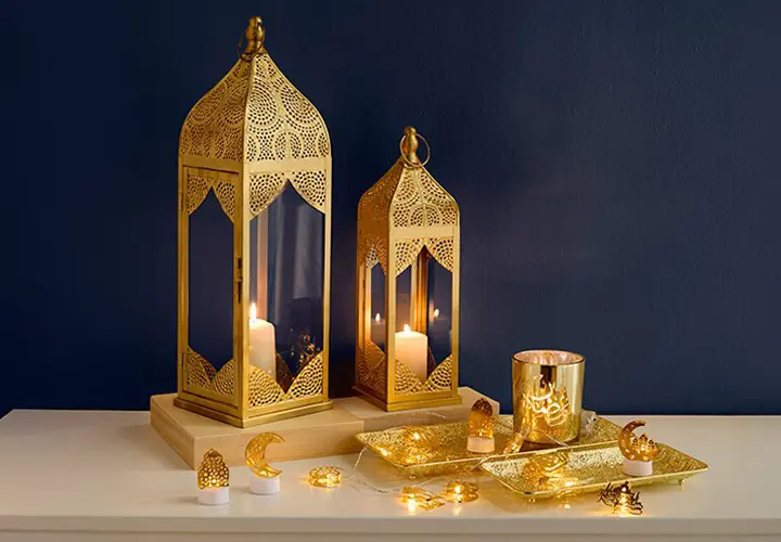 Filialverfügbarkeit Zwei verzierte goldene Laternen mit Kerzen, ein dekoratives Tablett und funkelnde Lichter vor einer dunkelblauen Wand.