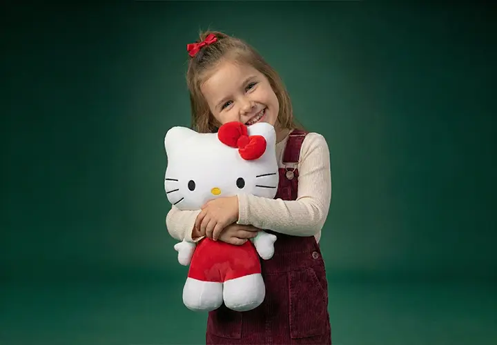 Ein Kind in einem bordeauxfarbenen Outfit umarmt ein großes Hello-Kitty-Plüschtier vor einem grünen Hintergrund. Ein Kind in einem bordeauxfarbenen Outfit umarmt ein großes Hello-Kitty-Plüschtier vor einem grünen Hintergrund.