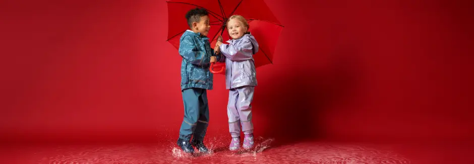 Zwei Kinder in bunten Regenmänteln planschen fröhlich in Pfützen, während sie sich einen leuchtend roten Regenschirm teilen. Zwei Kinder in bunten Regenmänteln planschen fröhlich in Pfützen, während sie sich einen leuchtend roten Regenschirm teilen.