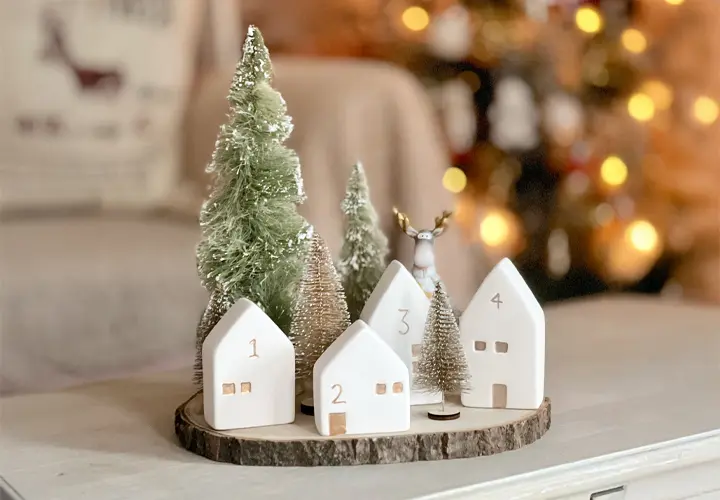 Eine verspielte Winterszene mit weißen Keramikhäusern nummeriert von 1 bis 4, grünen Miniaturbäumchen und einer Rentierfigur auf einer Holzplatte. Eine verspielte Winterszene mit weißen Keramikhäusern nummeriert von 1 bis 4, grünen Miniaturbäumchen und einer Rentierfigur auf einer Holzplatte.