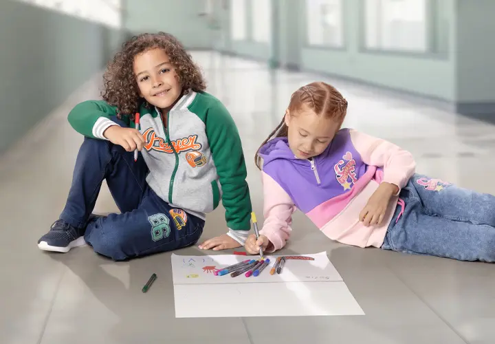 Zwei Kinder sitzen auf einem gefliesten Boden und malen mit farbigen Stiften auf einem großen Blatt Papier.