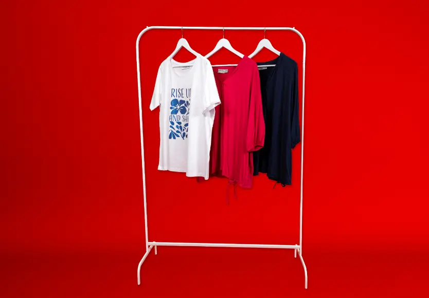 Drei stilvolle Oberteile hängen auf einem weißen Kleiderständer vor einem leuchtend roten Hintergrund: ein weißes T-Shirt mit Aufdruck, eine rosa Bluse und ein marineblaues Hemd. Drei stilvolle Oberteile hängen auf einem weißen Kleiderständer vor einem leuchtend roten Hintergrund: ein weißes T-Shirt mit Aufdruck, eine rosa Bluse und ein marineblaues Hemd.