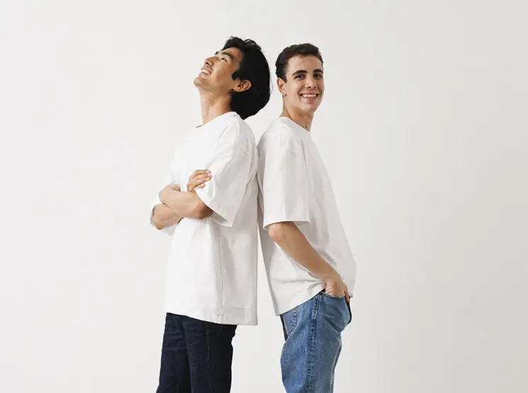 Zwei junge Männer in weißen T-Shirts stehen Rücken an Rücken vor einem schlichten weißen Hintergrund und lächeln entspannt.