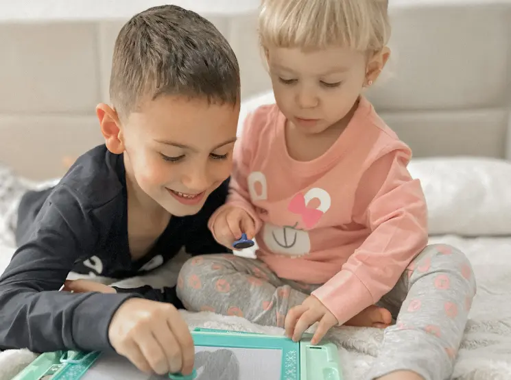 Zwei kleine Kinder sitzen auf einem Bett, spielen fröhlich miteinander und mit einem Zeichentablet, umgeben von weicher Bettwäsche.
