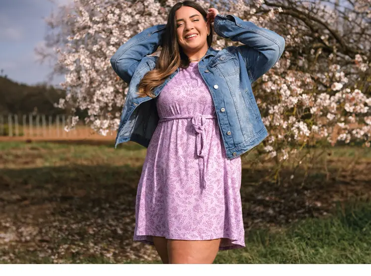 Eine Frau in einem lila Kleid und einer Jeansjacke posiert vor einem blühenden Baum.