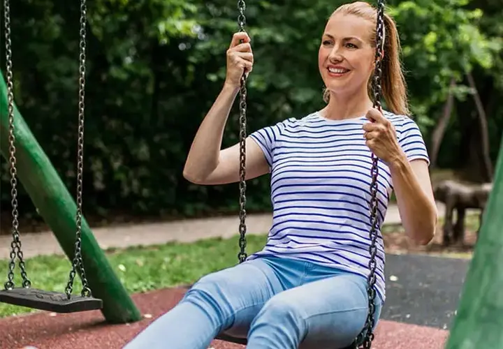 Eine Frau mit gestreiftem Shirt und Jeans schaukelt fröhlich in einem Park, umgeben von üppigem Grün.