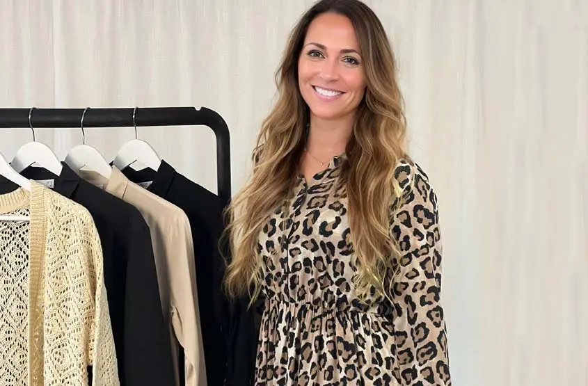 Eine Frau in einem Kleid mit Leopardenmuster steht neben einem Regal, auf dem verschiedene modische Kleidungsstücke vor einem neutralen Hintergrund ausgestellt sind.