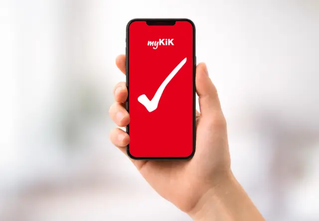 Eine Person hält ein Smartphone in der Hand, auf dem die myKiK-App mit einem roten Hintergrund und einem weißen Häkchen angezeigt wird.