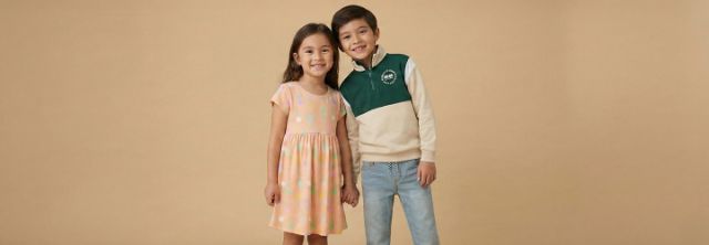 Zwei Kinder stehen zusammen vor einem schlichten beigen Hintergrund. Das Mädchen trägt ein buntes Kleid, während der Junge ein grün-cremefarbenes Sweatshirt trägt.