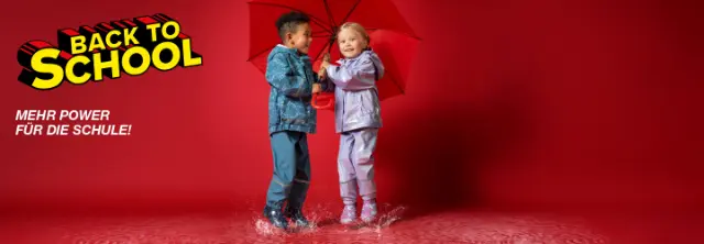 Zwei Kinder in bunten Regenmänteln planschen fröhlich in Pfützen, während sie sich einen leuchtend roten Regenschirm teilen.