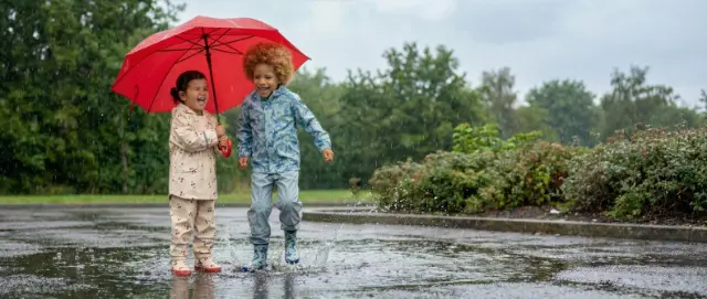 Zwei Kinder spielen im Regen, planschen in Pfützen und teilen sich einen knallroten Regenschirm, umgeben von viel Grün.