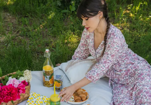 Eine Picknickszene mit einer Frau in einem geblümten Kleid, umgeben von bunten Blumen, einem Orangengetränk, Croissants und einer farbenfrohen Decke auf dem Rasen.