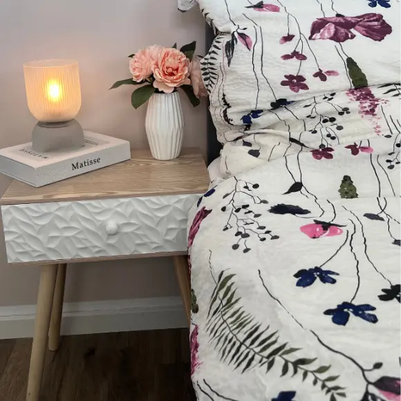 Eine gemütliche Nachttischszene mit einer leuchtenden Lampe, einer Blumenvase mit rosa Rosen und einer gemusterten Bettdecke vor einer sanften, neutralen Wand.