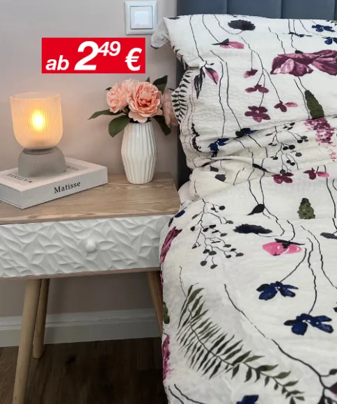 Eine gemütliche Nachttischszene mit einer leuchtenden Lampe, einer Blumenvase mit rosa Rosen und einer gemusterten Bettdecke vor einer sanften, neutralen Wand.