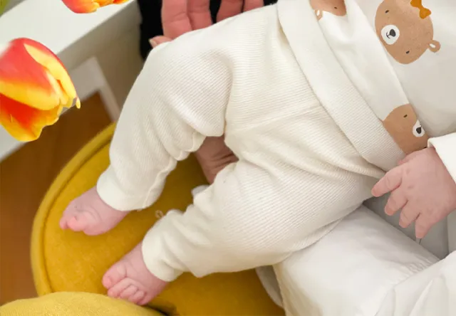 Eine Nahaufnahme der Beine eines Babys in einem Schlafanzug mit Bärenmuster, das auf einem Schoß liegt, mit leuchtenden Tulpen im Vordergrund.