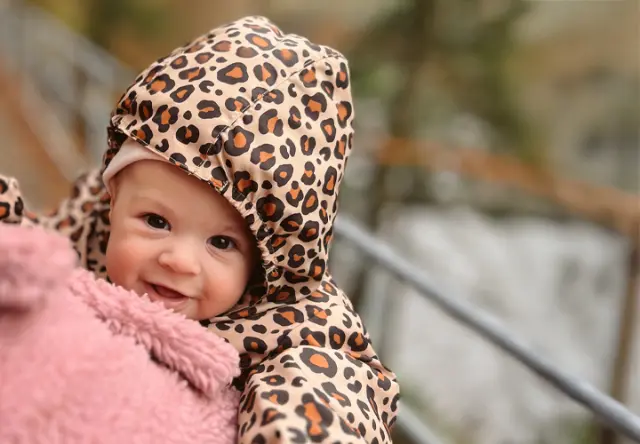 Ein Baby in einer Kapuzenjacke mit Leopardenmuster kuschelt sich in eine weiche rosafarbene Decke vor einer natürlichen Kulisse.
