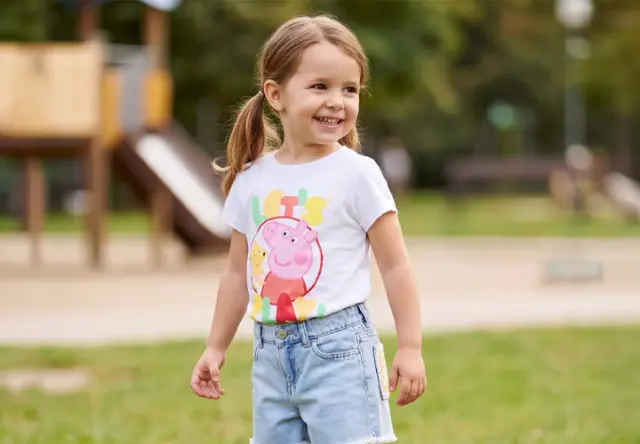 Mädchen mit Peppa-Wutz-T-Shirt und Jeansshorts auf einem Spielplatz – fröhliche Kinderkleidung für aktive Sommertage