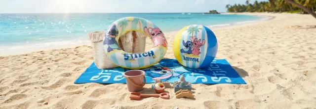 Strandtuch, Schwimmring, Ball und Sandspielzeug mit Lilo‑&amp;‑Stitch‑Motiv liegen am Sandstrand neben türkisfarbenem Meer.